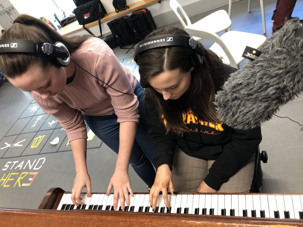 Two people with headphones on being recorded playing a piano.