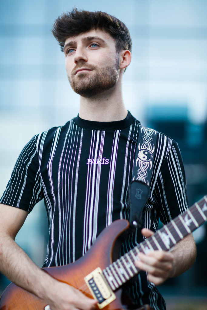 Person with beard and short quiffed hair, staring to the distance holding a guitar.