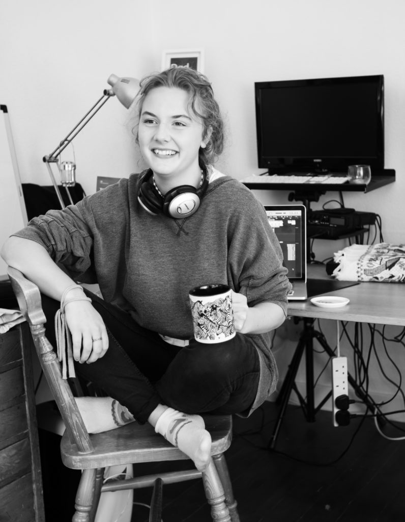 Young woman sitting crossed legged on a chair, holding a mug and smiling. Wearing headphones and in front of home studio.