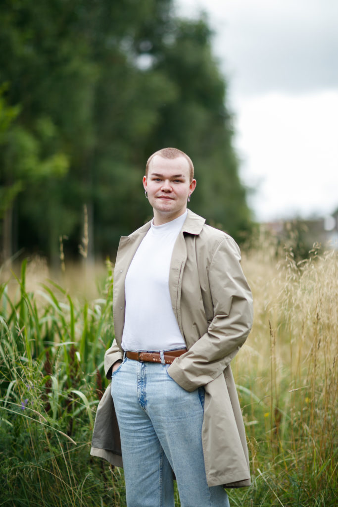 Person standing in field, hands in pockets smiling at camera. Skinhead with hoop earrings and trench coat.