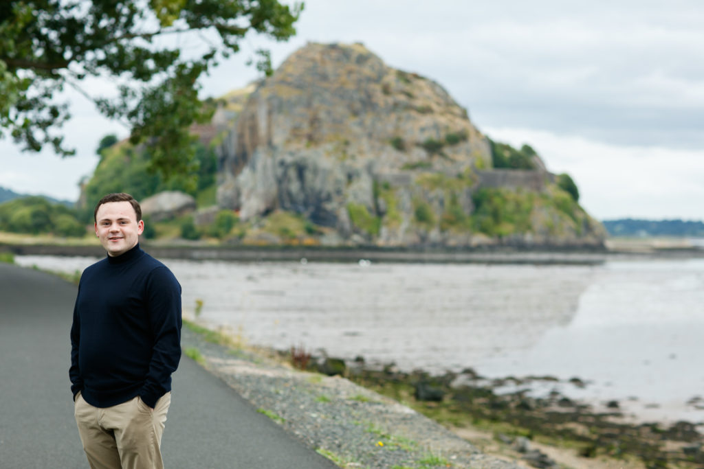 Young man standing in front of body of water with large rockface at the back.