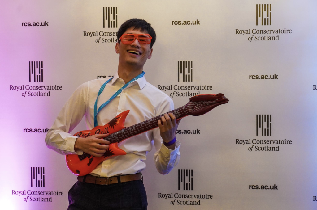 Young man in front of RCS banner, wearing prop glasses and blow up guitar