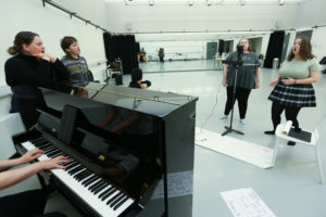 Young people in drama studio standing around piano
